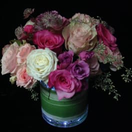 Compact arrangement of pink, peach, and cream roses in a clear glass vase with a green leaf wrap.