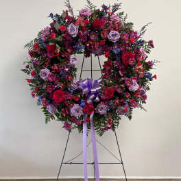 Large floral wreath on a stand with pink, purple, and blue flowers and a lavender ribbon