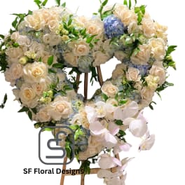 Heart-shaped standing arrangement of cream roses, blue hydrangeas, and cascading white orchids on a wooden easel.