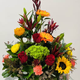 Mixed bouquet with red roses, orange gerberas, yellow blooms, and green hydrangea in a square vase