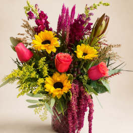 Mixed bouquet with sunflowers, roses, and magenta spikes in a glass vase