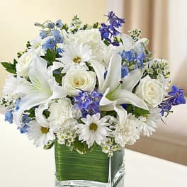White lilies, roses, and daisies with blue flowers in a square glass vase