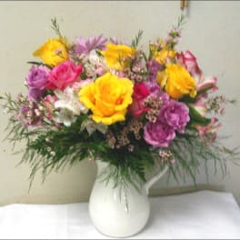 Mixed bouquet of yellow roses with pink and purple flowers in a white ceramic pitcher