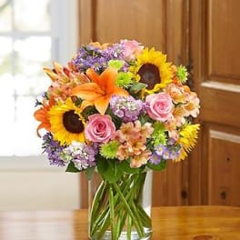 Mixed bouquet of sunflowers, roses, lilies, and purple flowers in a glass vase