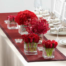 Red floral centerpieces in clear square vases on a table runner