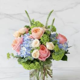 Low arrangement of peach and pink roses, cream roses, and blue hydrangeas in a clear glass vase.