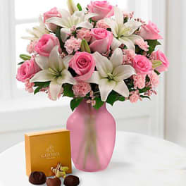 Pink roses and white lilies in a pink vase with a box of chocolates