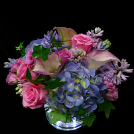 Low arrangement of pink roses, blue hydrangeas, and pale calla lilies in a clear glass vase.
