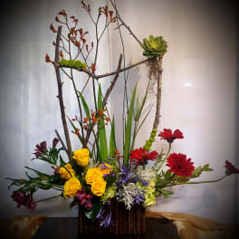 Tall floral arrangement with yellow roses, red daisies, and branches in a dark vase