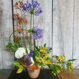 Tall mixed floral arrangement in a beige vase with purple, yellow, white, and orange blooms.