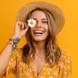 Smiling woman in a straw hat holding a white daisy over one eye