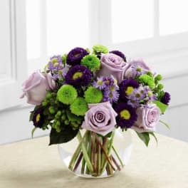 Lavender roses with purple asters and green button mums in a round clear glass vase