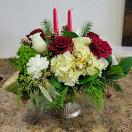 Holiday centerpiece with red roses, white hydrangeas, evergreens, and two red taper candles in a silver pedestal bowl