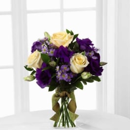 Cream roses with purple blooms and lavender asters in a clear vase tied with a green ribbon