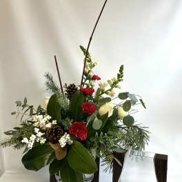 Holiday floral arrangement with red carnations, white blooms, pinecones, and greenery in a glass vase
