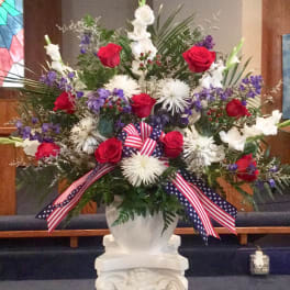 Tall red, white, and purple flower arrangement in a white urn with patriotic ribbon on a pedestal