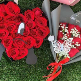 Heart-shaped box of red roses beside a wrapped bouquet of red roses