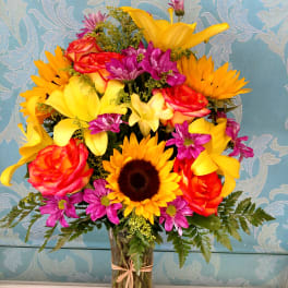 Bright mixed bouquet in a clear glass vase with sunflowers, roses, and purple daisies