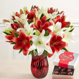 Red and white lilies in a red glass vase beside an open box of chocolates