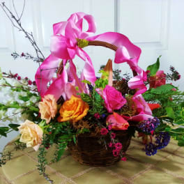 Basket arrangement with roses and mixed flowers topped by a pink ribbon bow