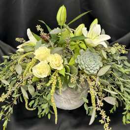 White lilies and pale roses in a low arrangement with trailing greenery and a succulent.