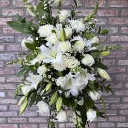 Tall all-white standing spray with lilies, roses, and mums on a metal easel against a brick wall.