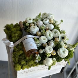 White flowers and chocolates arranged around a wine bottle in a moss-covered box