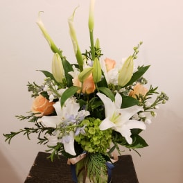 Tall arrangement of white lilies and peach roses with green hydrangea in a clear glass vase