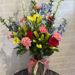 Tall mixed bouquet of yellow lilies, red roses, pink carnations, and blue delphinium in a glass vase with ribbon