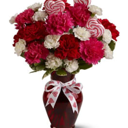 Red, pink, and white carnations in a red vase with a heart ribbon