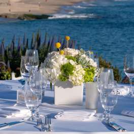 White floral centerpiece in a square vase on a set table by the water