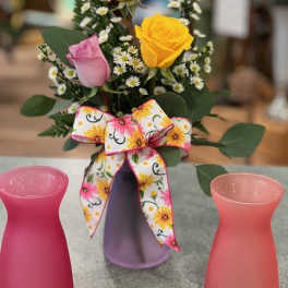 Small bouquet of roses and white filler flowers in a purple vase with a floral bow.