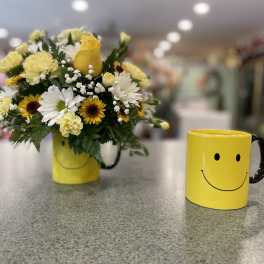 Yellow and white flower arrangement in a smiley-face mug beside a matching mug