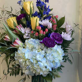 Tall mixed bouquet in a glass vase with blue, yellow, pink, and white flowers