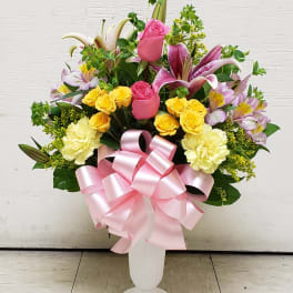 Tall arrangement of pink roses, lilies, yellow spray roses and carnations in a white vase with a pink bow
