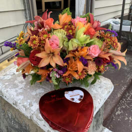Bright mixed bouquet with roses, lilies, and chrysanthemums beside a heart-shaped chocolate box.