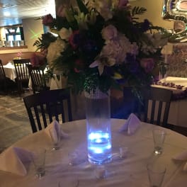 Tall floral centerpiece with pink, white, and purple blooms in a lit glass vase