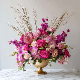 Tall arrangement of bright pink roses, orchids, and mixed blooms in a gold pedestal bowl