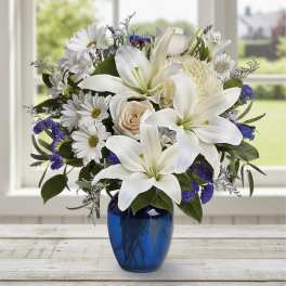 White lilies, daisies, and roses with purple accents arranged in a blue glass vase
