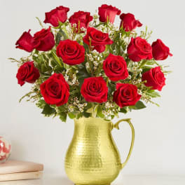 Arrangement of red roses with small white blooms in a hammered gold metal pitcher.