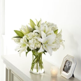 White lilies and hydrangeas in a clear glass vase on a table.