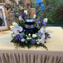 Cremation urn surrounded by blue hydrangeas, white roses, orchids and purple flowers on a draped table