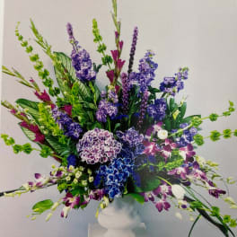 Tall arrangement of purple and blue hydrangeas, stock, orchids, and bells of Ireland in a white pedestal vase