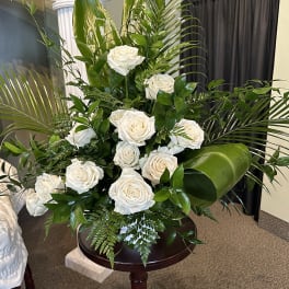 Tall arrangement of white roses with large green tropical leaves on a wooden pedestal.