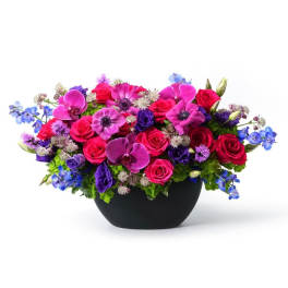 Low arrangement of hot pink roses, purple orchids, and blue flowers in a black bowl vase