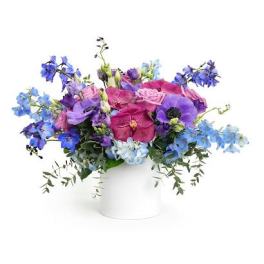 Low arrangement of pink roses and purple and blue blooms in a white cylinder vase