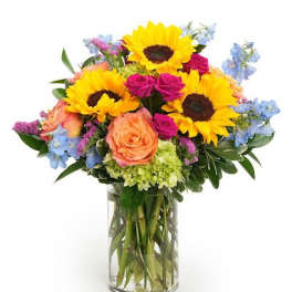 Arrangement of yellow sunflowers, pink roses, blue delphinium, and green hydrangeas in a clear glass vase.