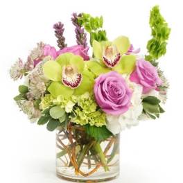 Short arrangement of green orchids, pink roses, and white blooms in a clear glass cylinder vase