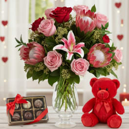 Pink and red bouquet with lilies and protea in a glass vase beside chocolates and a red teddy bear