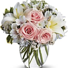 Round glass vase arrangement with pale pink roses, white lilies, and white accent flowers.
